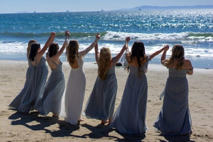 画像：ブライズメイドと手を繋ぎ両手を上げる花嫁が波打ち際に向かって砂浜を歩いている
