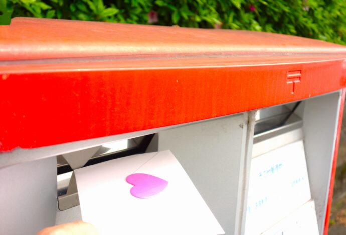画像：ピンクのハートで綴じた手紙を郵便ポストに投函しようとしているところ