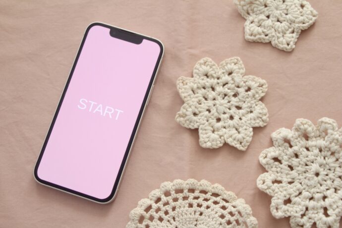 画像：ピンクの画面のスマートフォンと茶色いレース編みのコースター